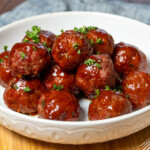 Smoked barbecue meatballs coated in a glossy, sweet, and smoky BBQ sauce, garnished with fresh parsley, served in a white bowl on a wooden board with toothpicks on the side.