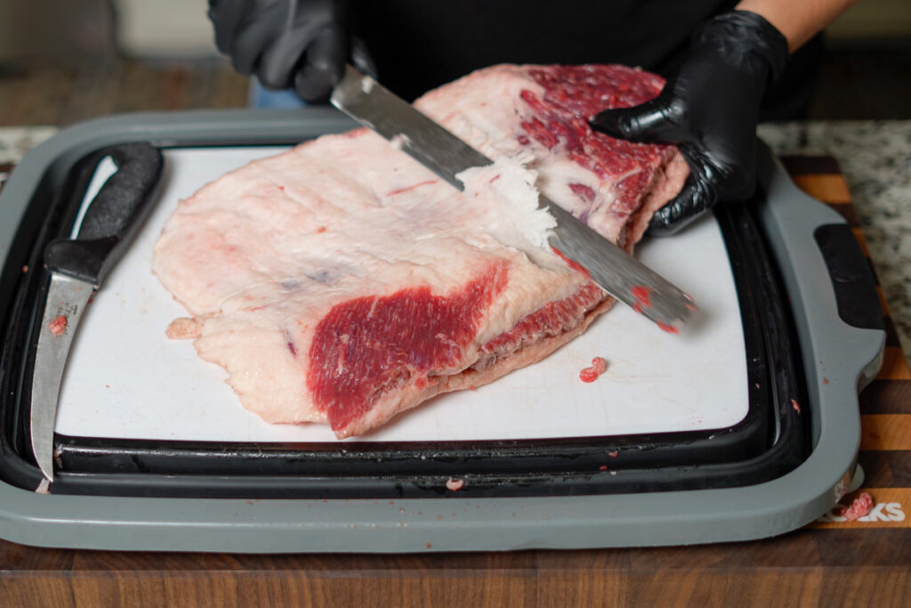 "A person wearing black gloves trims the fat from a raw brisket point on a white cutting board. The brisket has a thick fat cap, and a sharp knife is being used to remove excess fat for even cooking. A second knife rests on the cutting board beside the brisket. The setup is on a wooden countertop with a black and gray cutting board frame.