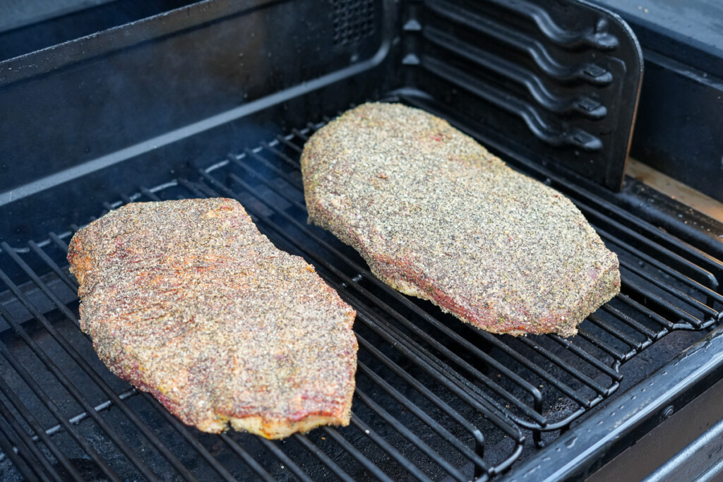 Two brisket cuts, the point and the flat, are placed separately on a smoker grate, seasoned with a coarse salt and pepper rub.