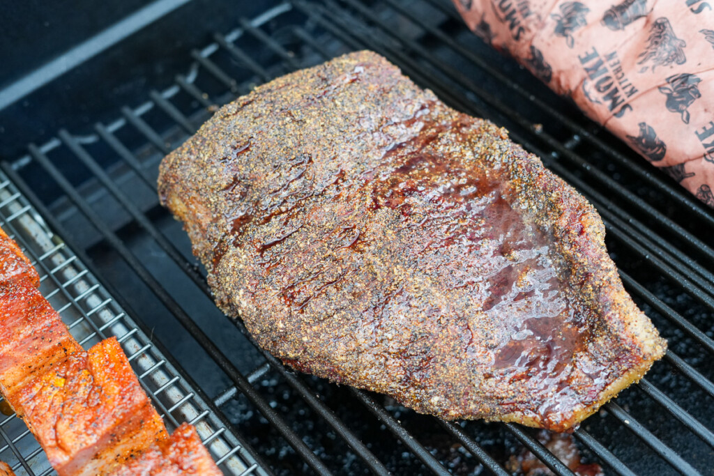 A brisket point on a smoker grate with a glistening bark, alongside a wrapped brisket flat and a wire rack holding seasoned meat.