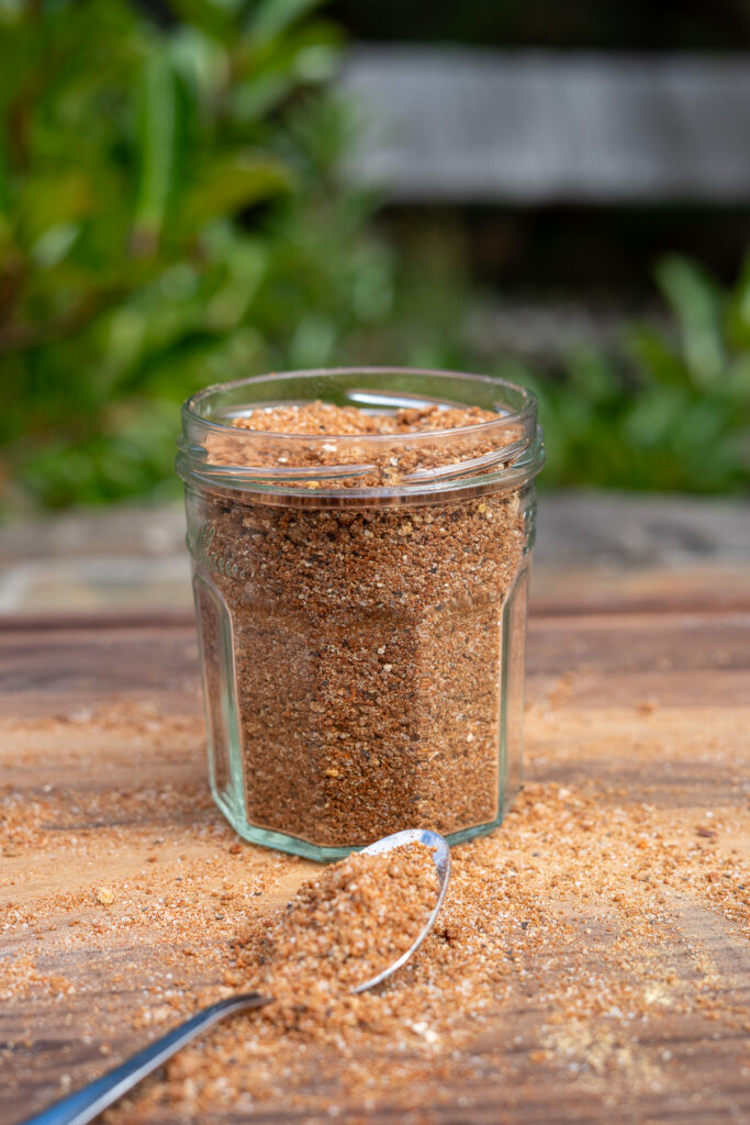 Glass jar filled with BBQ pork rub on a wooden surface, with a spoonful of the seasoning spilled in front.