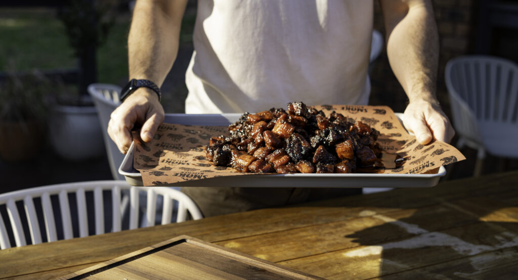 Person holding a tray of smoked pork belly burnt ends, glistening with barbecue glaze and served on branded butcher paper outdoors.