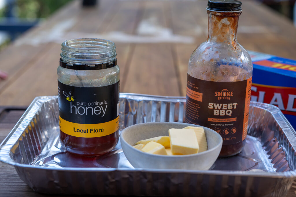 Aluminum tray containing a jar of honey, a bottle of sweet BBQ sauce, and a bowl of butter—ingredients for the pork belly burnt ends glaze.