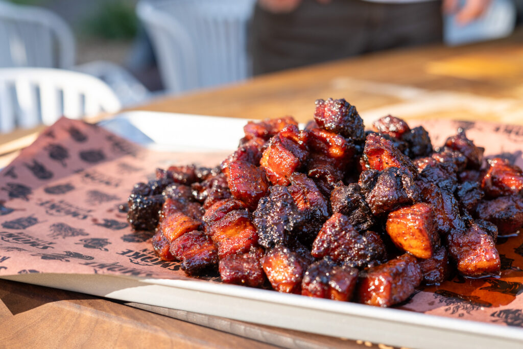 Close-up of glazed pork belly burnt ends piled on butcher paper, showing crispy bark and a sticky, caramelized BBQ glaze in natural sunlight.