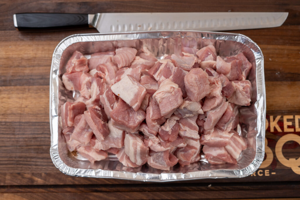 Tray filled with evenly cubed raw pork belly, ready for seasoning and smoking, with a slicing knife resting on a wooden cutting board.