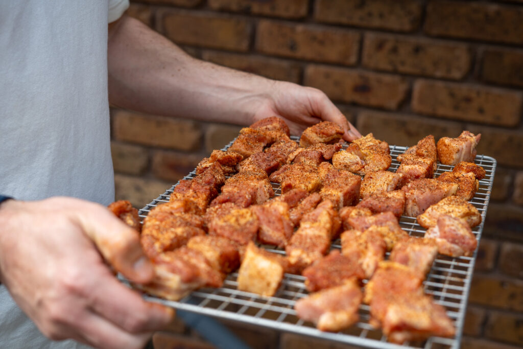 Person holding a wire rack loaded with seasoned pork belly cubes, ready to be placed in the smoker for burnt ends.