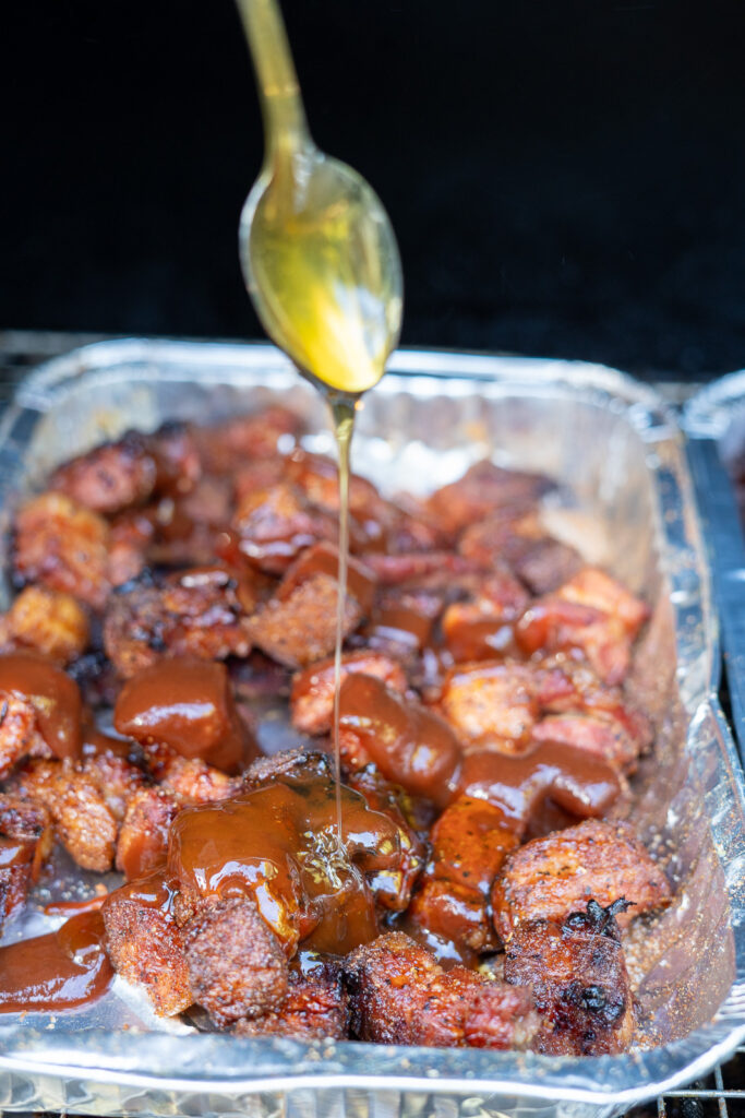 Honey being drizzled over smoked pork belly burnt ends in a foil tray, along with BBQ sauce, before covering and returning to the smoker to braise.