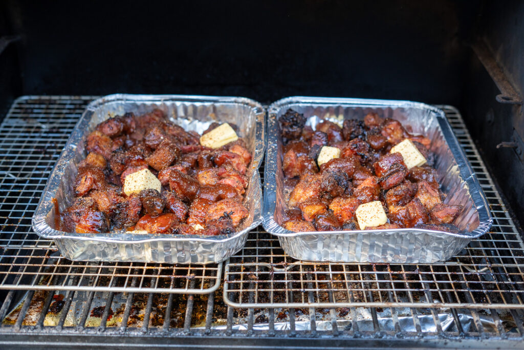 Two foil trays of pork belly burnt ends in a smoker, topped with BBQ sauce, honey, and pats of butter, ready to be covered and braised.