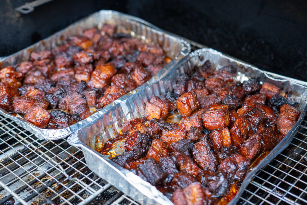 Two foil trays of pork belly burnt ends uncovered in the smoker, with a thick, caramelized glaze after the braising stage.