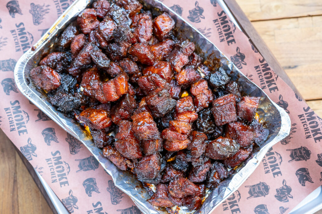 Overhead view of a foil tray filled with finished pork belly burnt ends, coated in a glossy barbecue glaze and served on branded butcher paper.