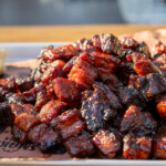 Close-up of glazed smoked pork belly burnt ends piled high on a tray with brioche buns and slaw, showcasing caramelized bark and sticky barbecue glaze.