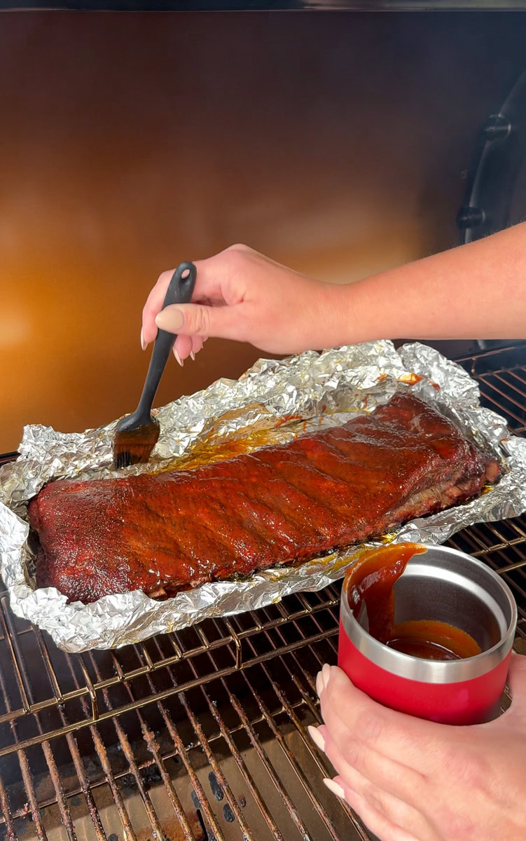 Person brushing barbecue sauce onto a rack of smoked ribs in foil on the smoker, with a red sauce cup in hand during the final stage of the 3-2-1 method.