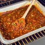 Smoked baked beans cooking in an aluminum pan on a smoker grate, stirred with a wooden spoon. Beans are thick, saucy, and flecked with diced jalapeño and onion.