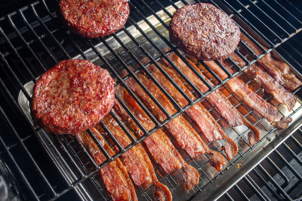 a tray of bacon cooking under thre burger patties on the smoker