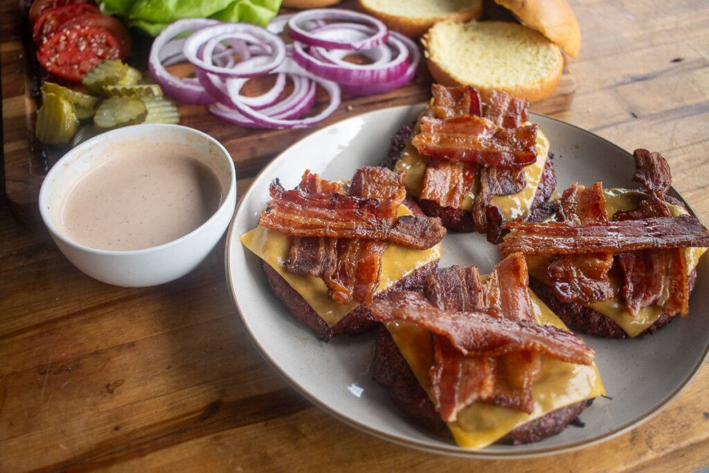 burger buns with a patty, cheese and bacon on it and a bowl of sauce