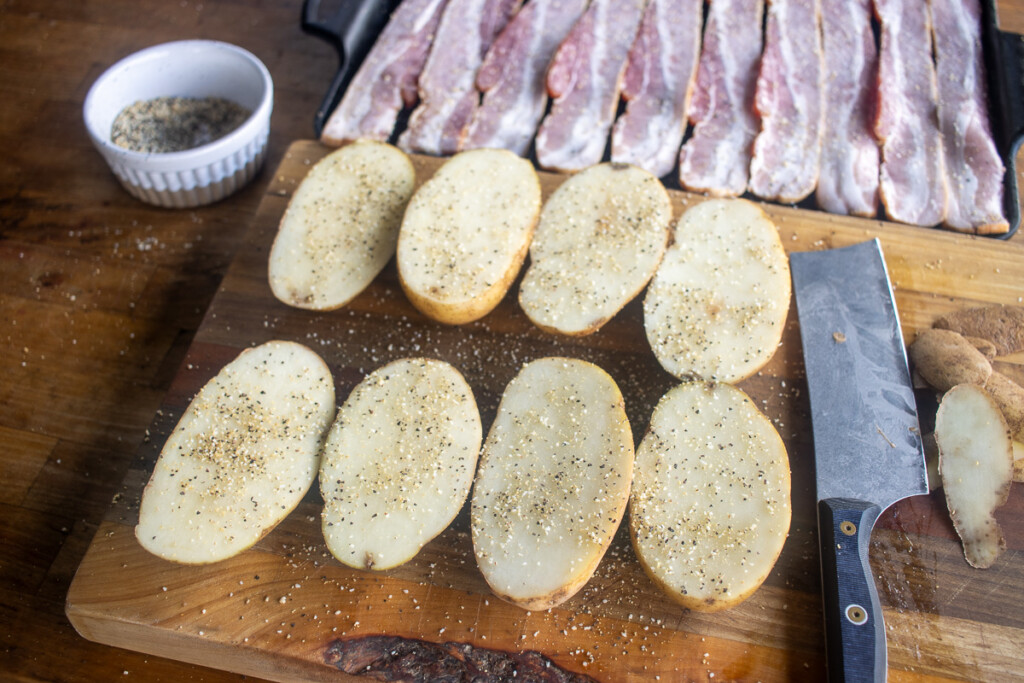 raw potatoes cut in half and sesoned on a wooden board with bacon in the background