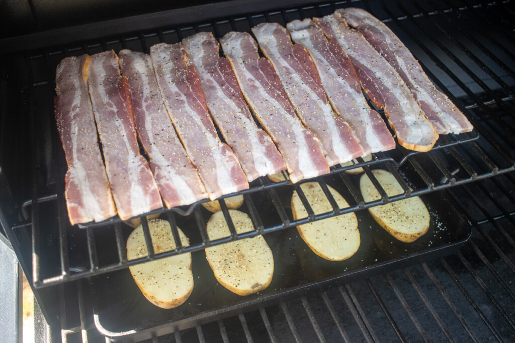 raw bacon strips on a rack above raw potato halves in the smoker