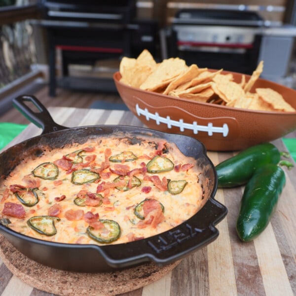 Jalapeno popper dip in a cast iron skillet with a football bowl filled with tortilla chips and two jalapenos.