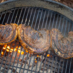 Three skewered picanha steaks grilling over charcoal on a round kettle grill, with visible flames and rich sear marks on the meat.