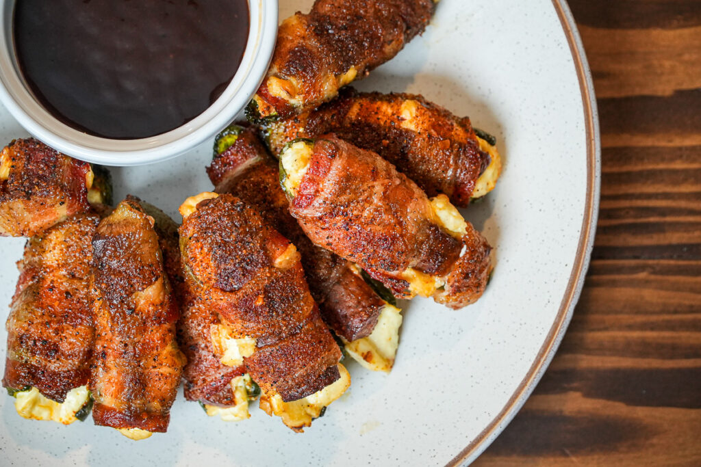 Plate of smoked Atomic Buffalo Turds—bacon-wrapped jalapeños stuffed with cream cheese and sausage—served with a side of barbecue sauce for dipping.
