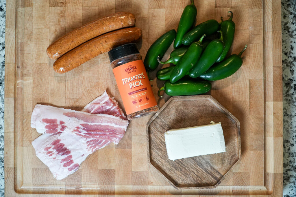 Ingredients for Atomic Buffalo Turds arranged on a wooden cutting board, including fresh jalapeños, cream cheese, bacon slices, smoked sausage links, and a bottle of Pitmaster’s Pick BBQ rub.