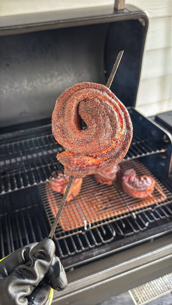 Gloved hand holding a smoked pork belly pinwheel on a skewer in front of a smoker, with more pinwheels cooking on a wire rack in the background.