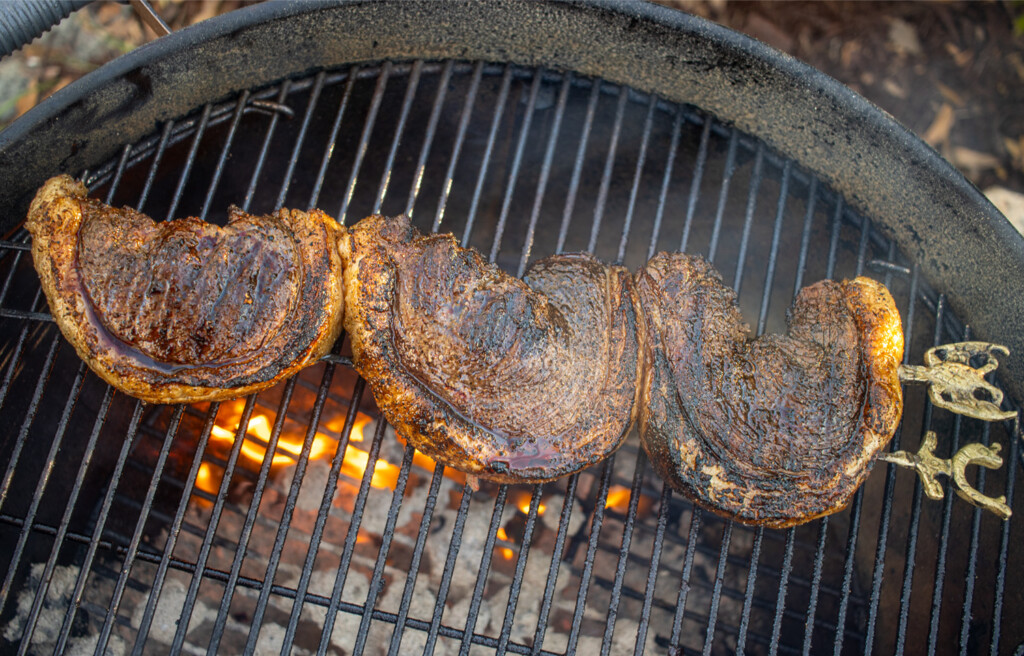 Three skewered picanha steaks grilling over charcoal on a round kettle grill, with visible flames and rich sear marks on the meat.