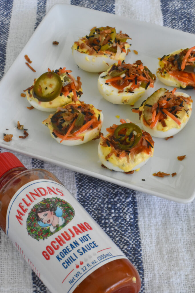 A plate of kimchi deviled eggs topped with jalapeño slices and shredded vegetables sits on a striped cloth next to a bottle of Melindas Gochujang Korean Hot Chili Sauce.