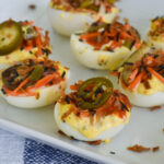 Six Kimchi Deviled Eggs on a white plate, topped with shredded carrots, crispy bits, and sliced jalapeños. The plate sits on a blue and white cloth.