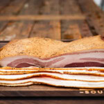 Slab of cured pork belly with sliced homemade bacon on a cutting board, showing marbling and peppered crust.