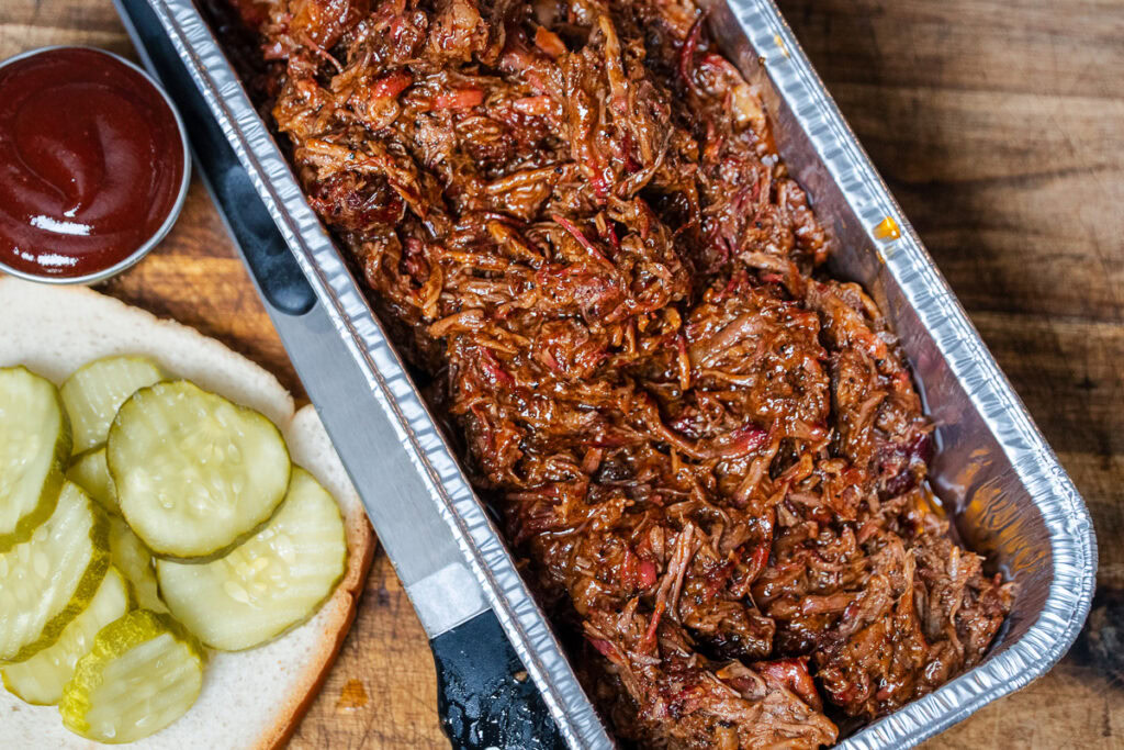 Shredded smoked chuck roast in a foil tray, ready to serve with white bread, barbecue sauce, and sliced pickles.