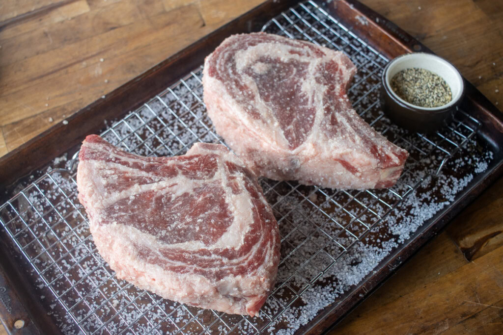two raw rib eye steaks on a wire rack with Kosher salt sprinkled all over them.