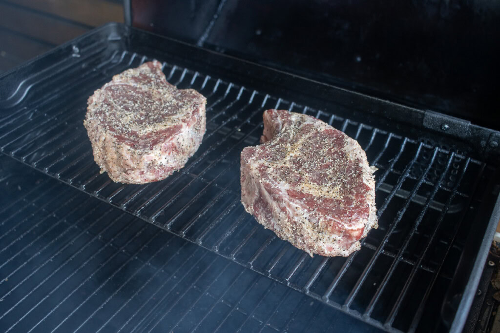 two raw seasoned rib eye steaks on the pellet smoker top rack
