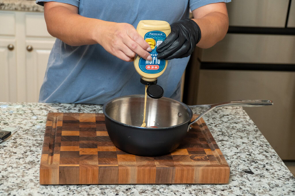 a person squeezing a bottle of Dijon into a metal bowl