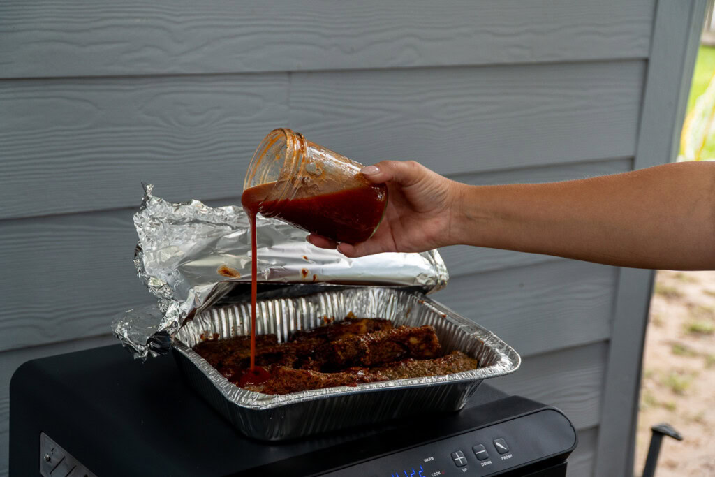 a person pouring maple bourbon bbq sauce over barbecued meat in a metal tray