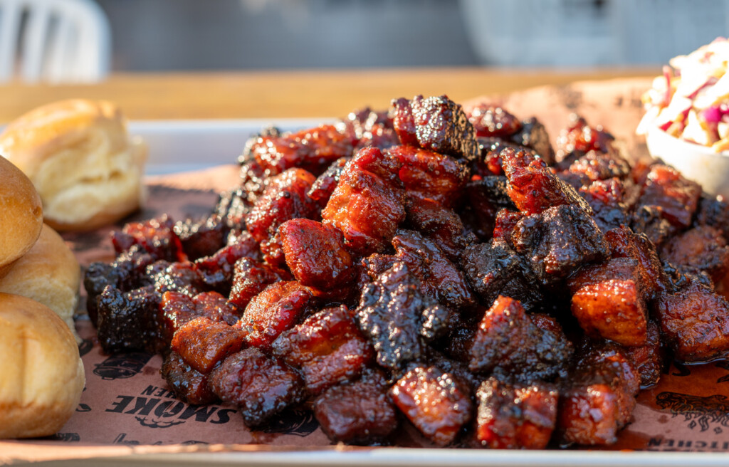Close-up of glazed smoked pork belly burnt ends piled high on a tray with brioche buns and slaw, showcasing caramelized bark and sticky barbecue glaze.