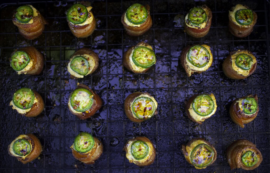 Overhead view of smoked pig shots on a wire rack, featuring crispy bacon-wrapped sausage cups filled with golden cream cheese, topped with sliced jalapeño and sweet BBQ seasoning.