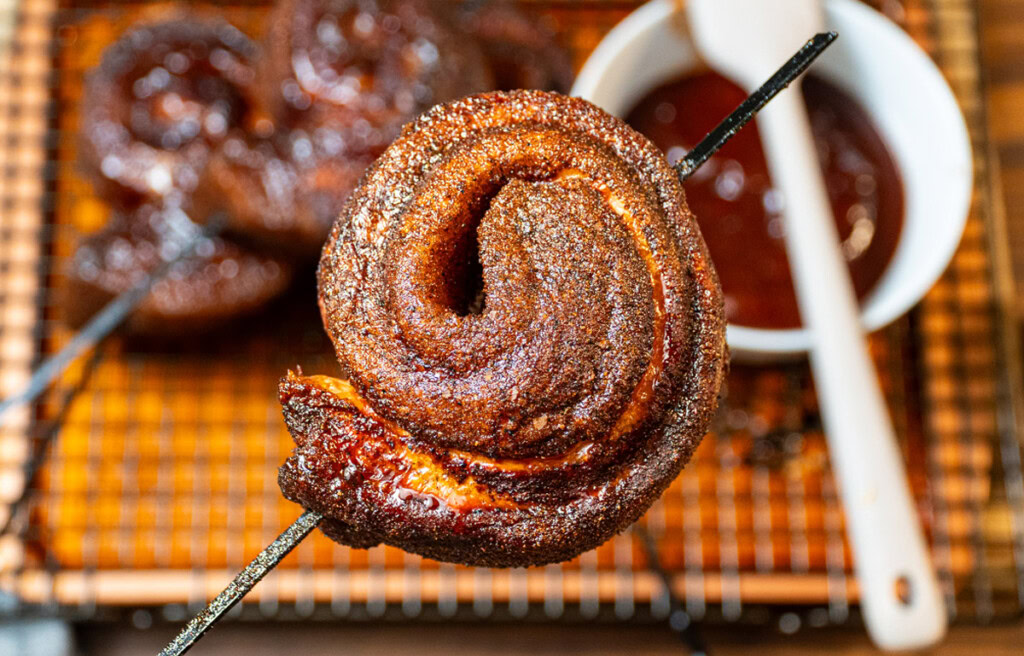 Close-up of a smoked pork belly pinwheel on a metal skewer, showing crispy bark and rendered fat, with barbecue sauce in the background.