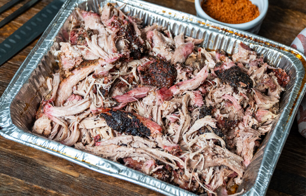 Tray of smoked pulled pork with dark bark and pink smoke ring, served in a foil pan on a wooden table with a bowl of dry rub in the background.