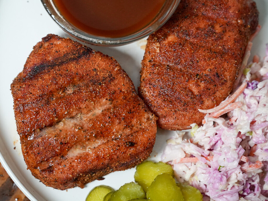 Two perfectly smoked pork chops with a deep, flavorful crust sit on a white plate, served with coleslaw, pickles, and a side of barbecue sauce. The grill marks and seasoning on the chops highlight their smoky, juicy goodness.