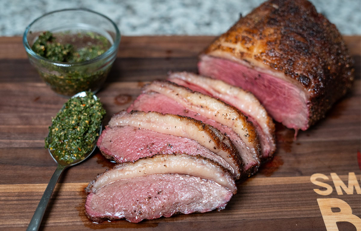 smoked and sliced picanha on a wood chopping board with a spoon full of chimichurri sauce