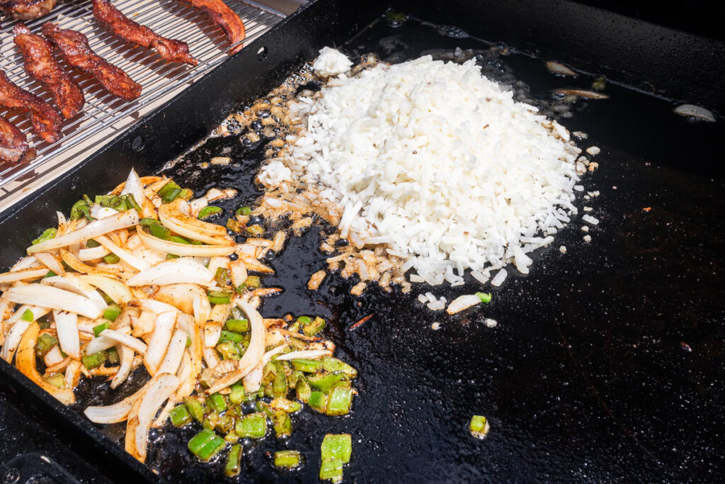a pile of uncooked shredded potato, and a pile of diced onion and green pepper on the griddle