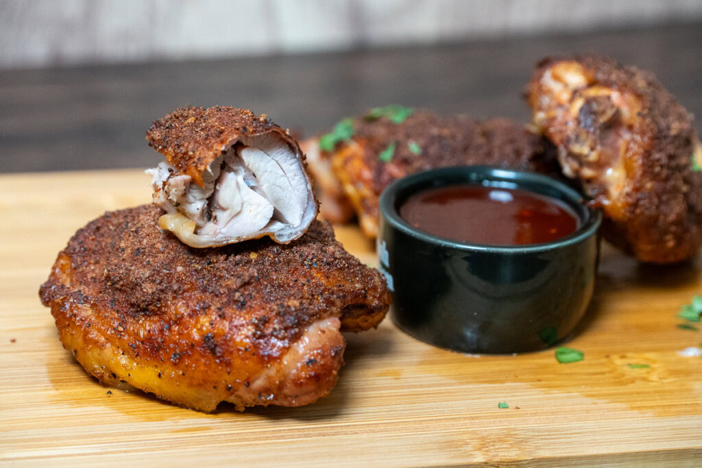 a cooked whole chicken thigh and a piece of thigh sitting on top showing the inside meat. a bowl of sauce on the side.