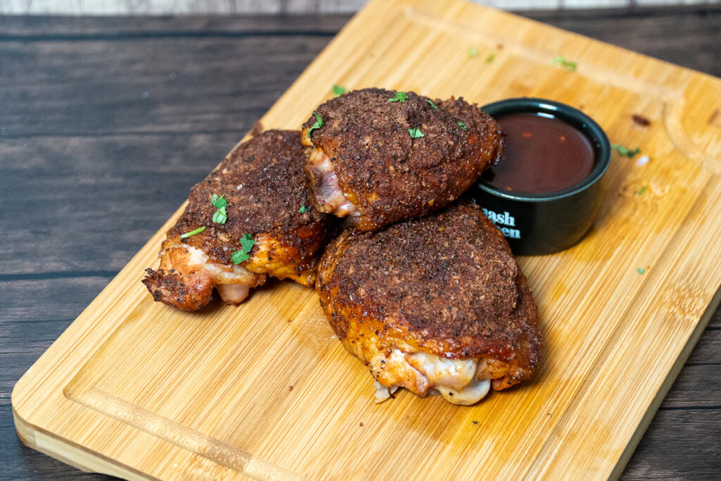 three pieces of cooked chicken thigh on a wooden board with a bowl of sauce