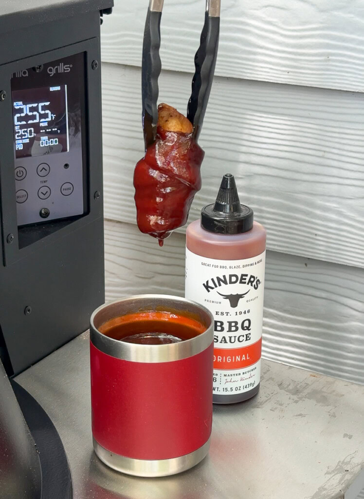 a pair of tongs holding a bacon wrapped chicken wing after dunking it in a tumbler full of BBQ sauce