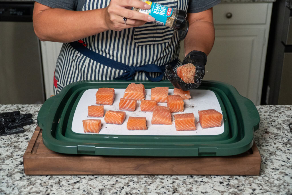a person sprinkling lemon pepper seasoning over cube up pieces of raw salmon