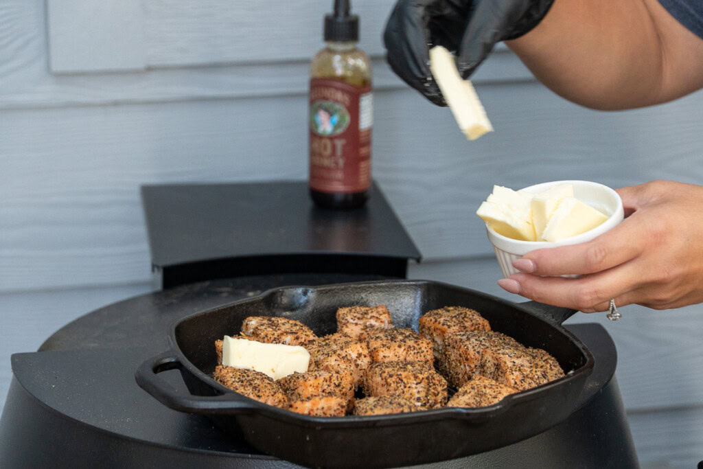 butter pats being added to a cast iron pan of cooked salmon cubes