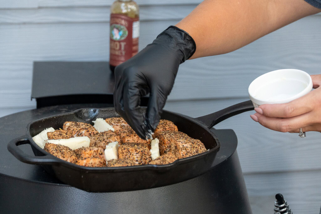butter pats being added to a cast iron pan of cooked salmon cubes