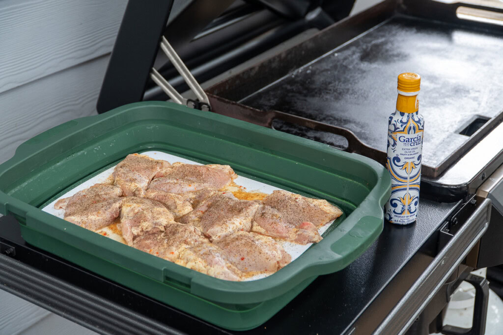 raw seasoned chicken in a tray sitting on the side of the griddle with a bottle of fancy oil