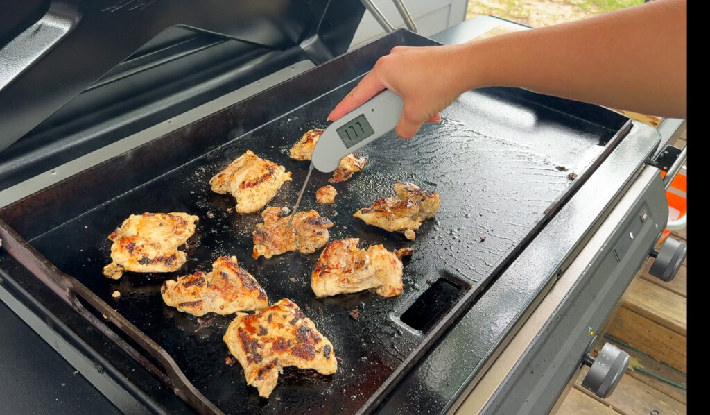 a hand holding a temperature probe inserted a chicken thigh on the griddle